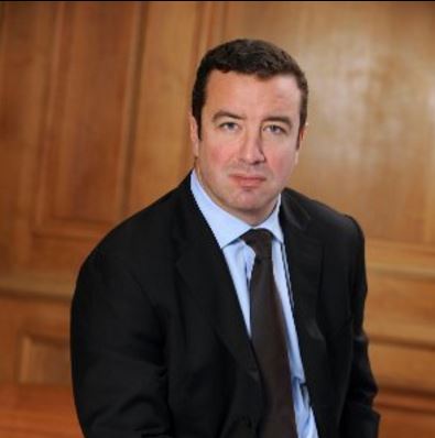 Man in a black suit and blue shirt sitting in front of a wooden wall.