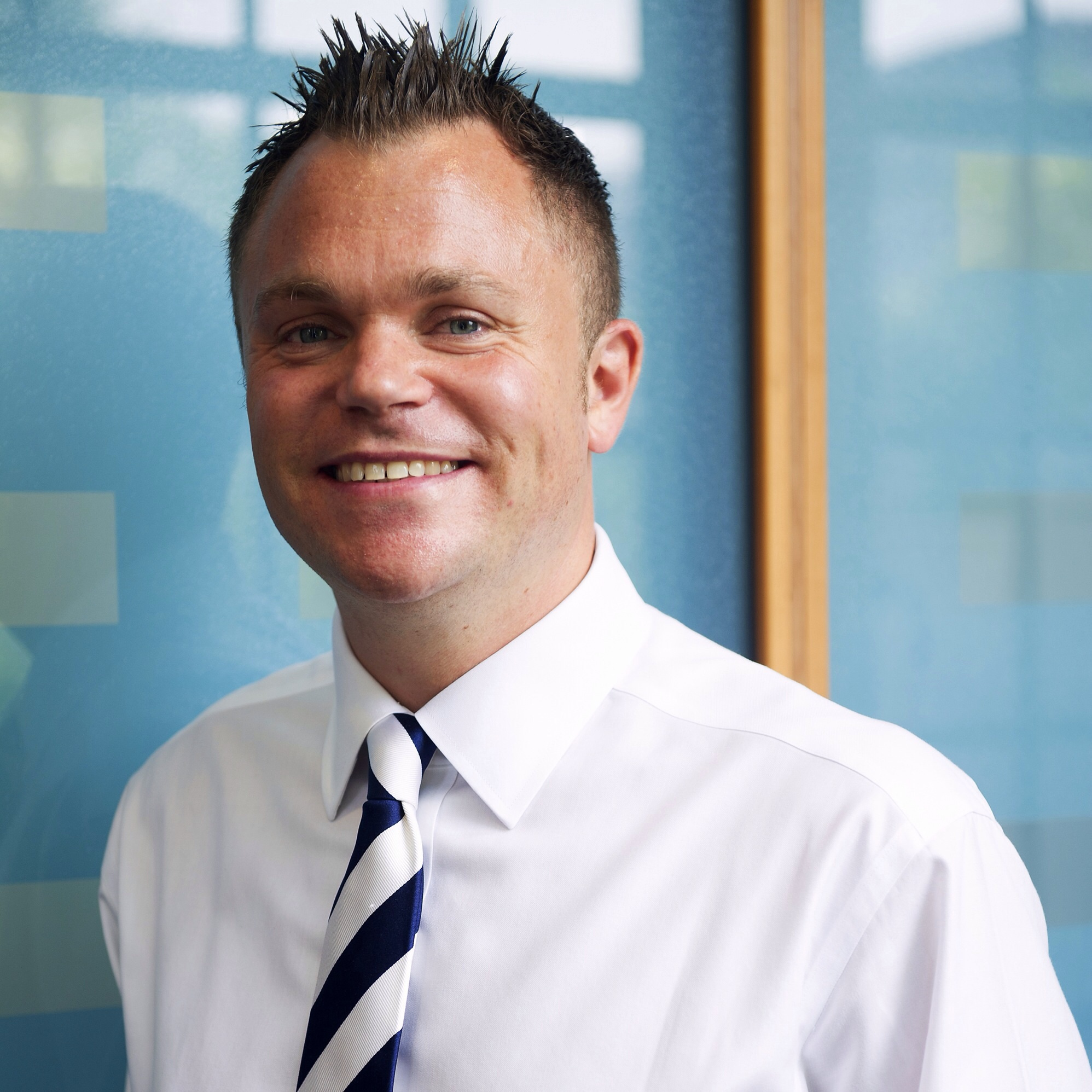 Smiling person in a white shirt with a striped tie against a blue background.