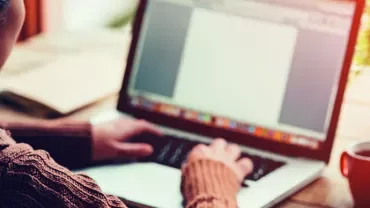 Person typing on a laptop at a desk with a red mug and plant.
