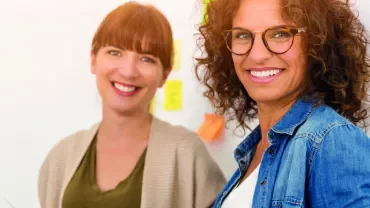 Two women smiling in an office with post-it notes on the wall.