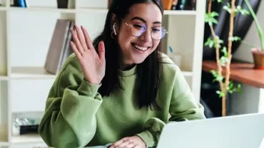 Woman in green sweater waving at laptop during video call.