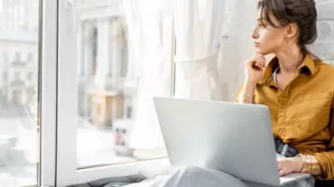 Woman with laptop gazing out the window thoughtfully.