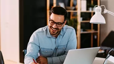 Man in a denim shirt working at a desk with a laptop.