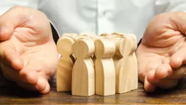 Hands surrounding wooden figures on a table, symbolizing protection.