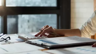 Person using calculator with papers and glasses on desk.