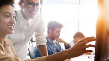 Two women in an office setting, looking at a computer screen and smiling.