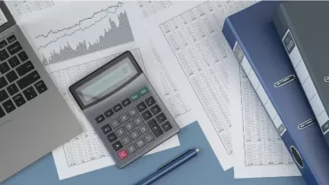 Laptop, calculator, and documents with charts and binders on a desk.