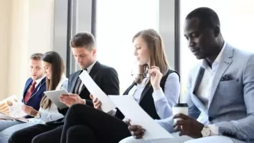 Business people seated in a waiting area, reviewing documents and tablets.