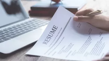 Person holding a resume near a laptop on a wooden table.