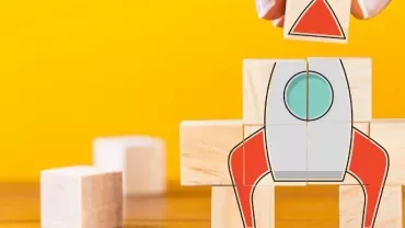 Stacked blocks forming a rocket with hand placing final piece, against a yellow background.