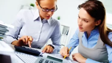 Two women reviewing documents and using a calculator at a desk.