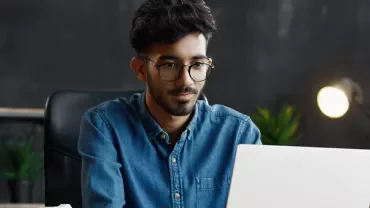 Man in glasses working on a laptop at a desk with plants and a lamp.