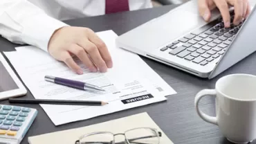 Person working on a laptop with documents, a pen, and glasses on the desk.