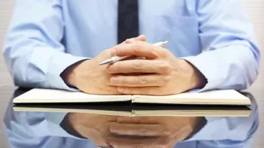 Person in a blue shirt with hands clasped over an open notebook on a reflective table.