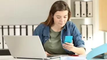 Person using a smartphone while working on a laptop at a desk.