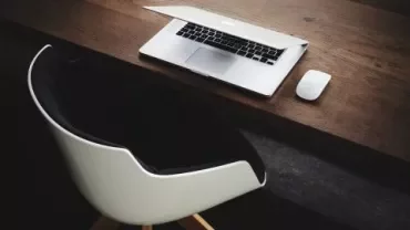 Open laptop and mouse on a wooden desk with a black and white chair.