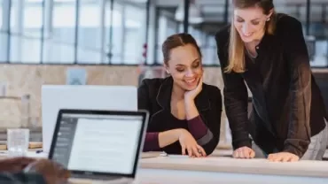 Two women smiling and discussing work in a modern office setting.