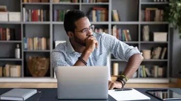 Man in an office thinking while sitting at a desk with a laptop.