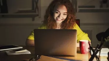 Woman with curly hair smiling at laptop screen in dim room.
