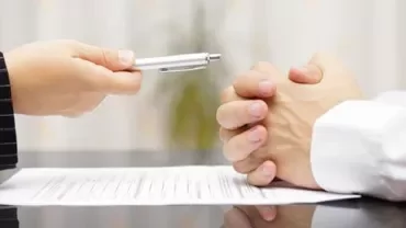 Person handing a pen over a document to another person at a desk.
