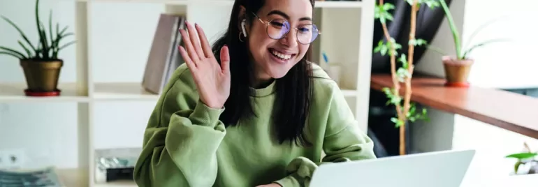 Woman in green sweater waving at laptop during video call.