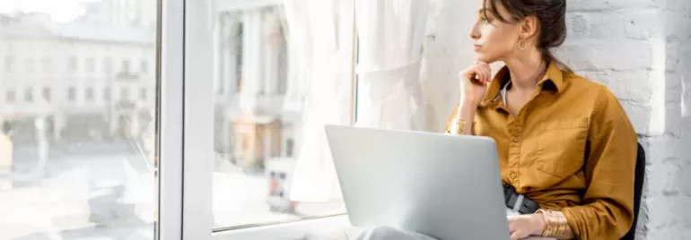 Woman with laptop gazing out the window thoughtfully.