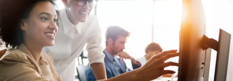 Two women in an office setting, looking at a computer screen and smiling.