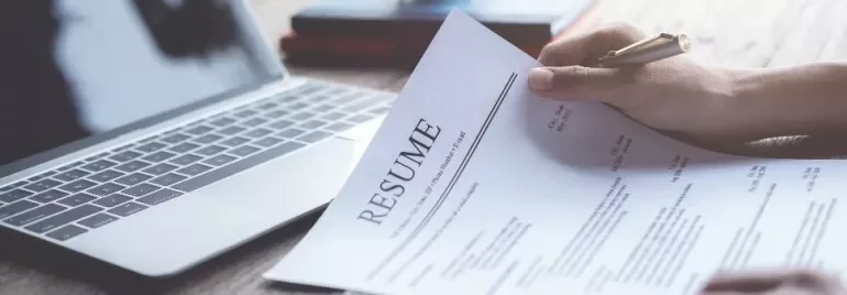 Person holding a resume near a laptop on a wooden table.