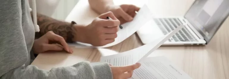 Two people reviewing documents at a desk with a laptop.