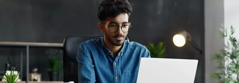 Homme avec des lunettes travaillant sur un ordinateur portable à un bureau avec des plantes et une lampe.