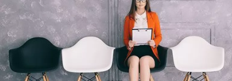 Woman in orange blazer sitting in a chair holding a clipboard.