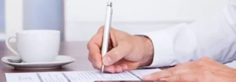 Person writing on a paper beside a cup and smartphone.