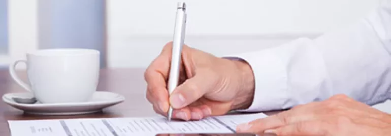 Person signing document at a desk with a cup of coffee and a phone.