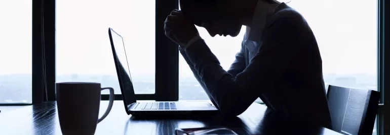 Silhouette of person stressed over laptop at desk with coffee mug.