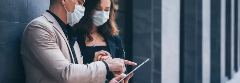 Two people wearing masks, discussing something on a tablet.