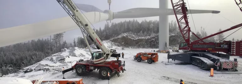 Workers installing a wind turbine blade with cranes in a snowy landscape.