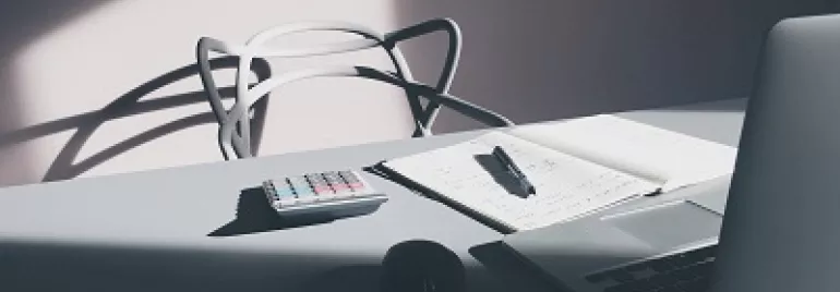 Un ordinateur portable, carnet, stylo et calculatrice sur un bureau ensoleillé.