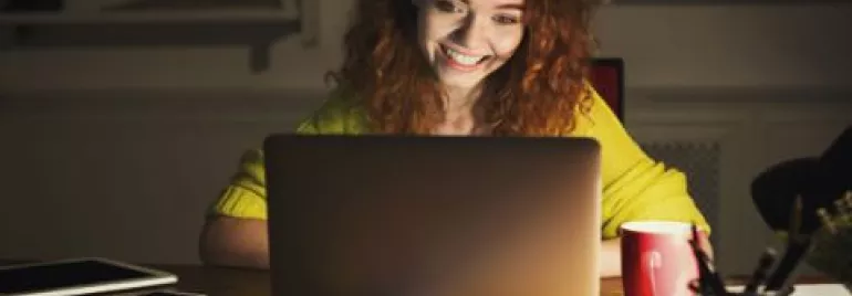 Woman with curly hair smiling at laptop screen in dim room.