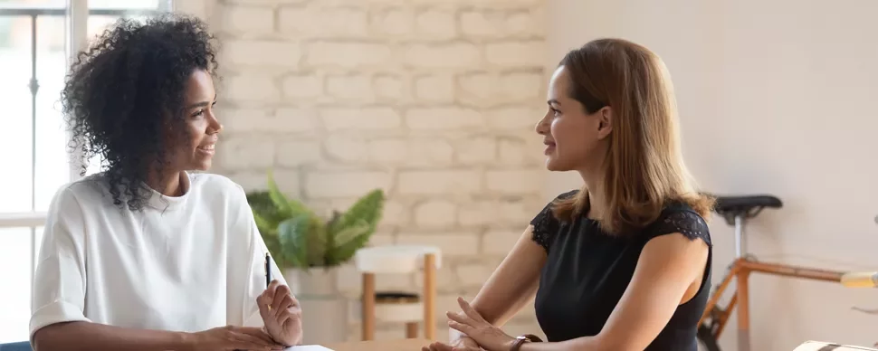 Two women having a discussion at a table with a brick wall in the background.