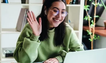 Woman in green sweater waving at laptop during video call.