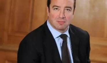 Man in a suit sitting against a wooden paneled background.