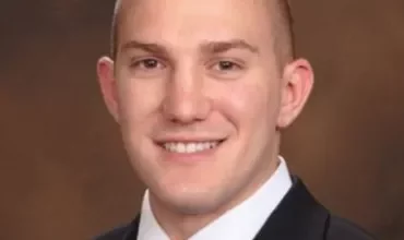 A person in a suit and tie smiling against a brown background.