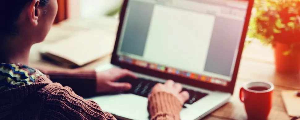 Person typing on a laptop at a desk with a red mug and plant.