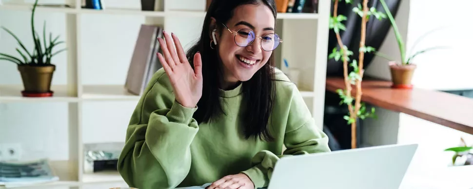 Woman in green sweater waving at laptop during video call.