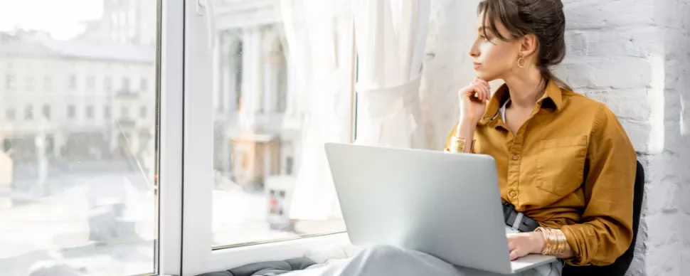 Woman with laptop gazing out the window thoughtfully.