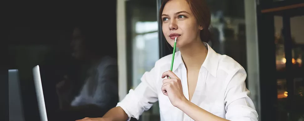 Woman thinking at desk with laptop, holding pencil.