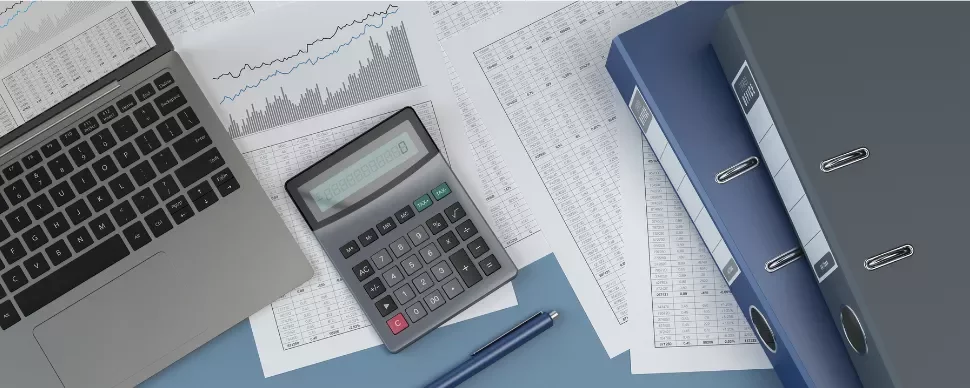 Laptop, calculator, and documents with charts and binders on a desk.
