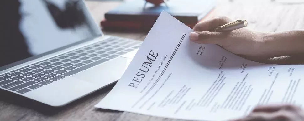 Person holding a resume near a laptop on a wooden table.