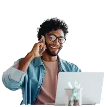 A person seated at a desk using a laptop, holding a phone to their ear, with a small desk accessory in front.