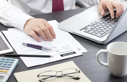 Person typing on laptop with documents, glasses, and a calculator on the desk.